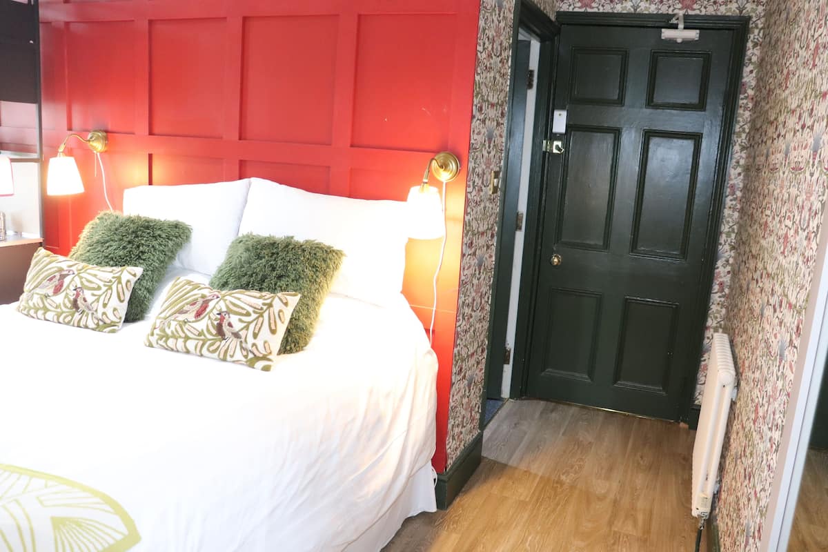 A hotel room with a red wall, a bed with white bedding and green pillows, a door with a floral wallpaper, and a radiator.
