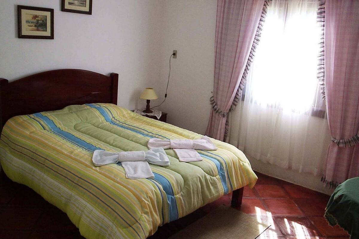 Una cama bien hecha con ropa de cama y toallas a rayas, una lámpara de noche y una ventana con cortinas.
