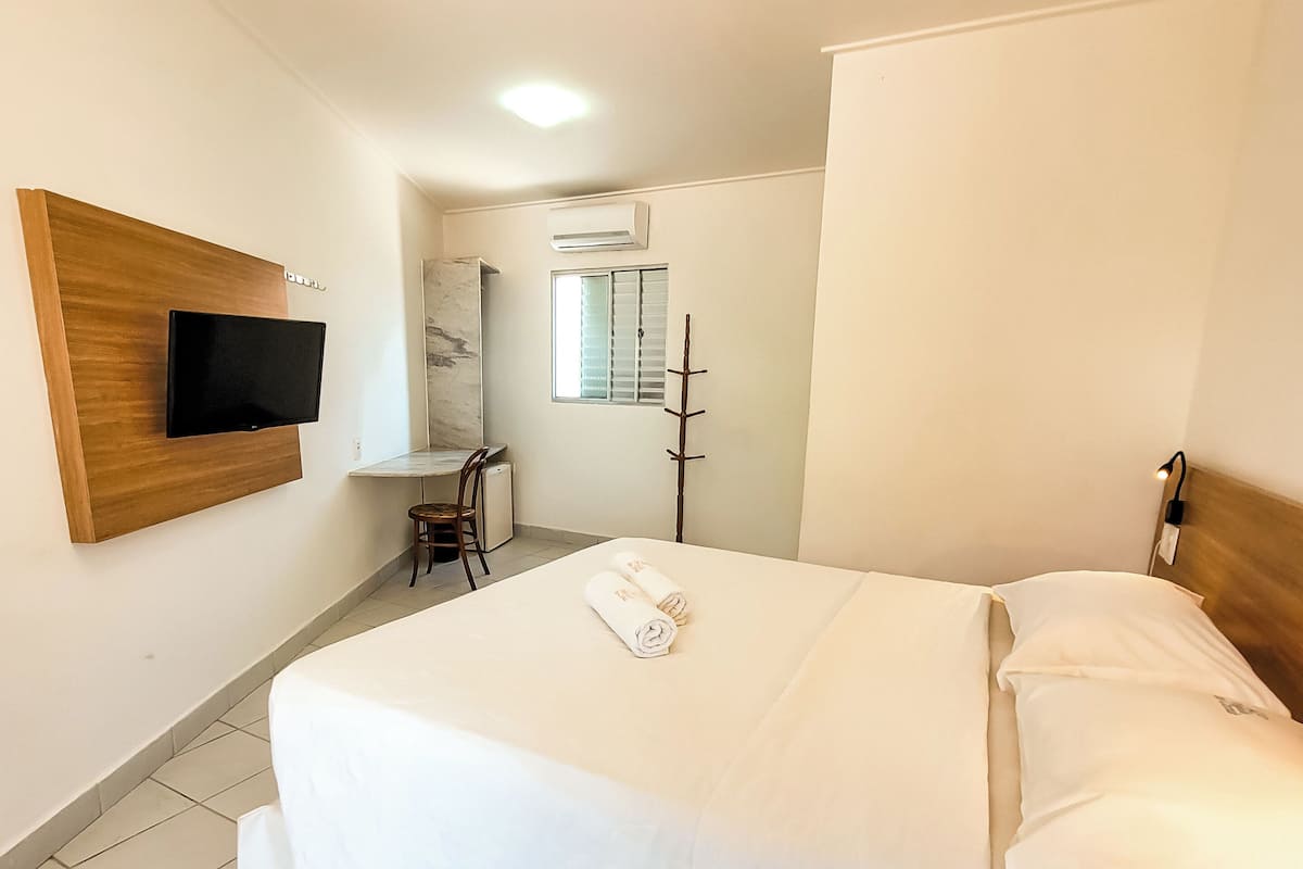 Una habitación de hotel con cama, televisión montada en la pared, escritorio con silla y ventana con persianas.