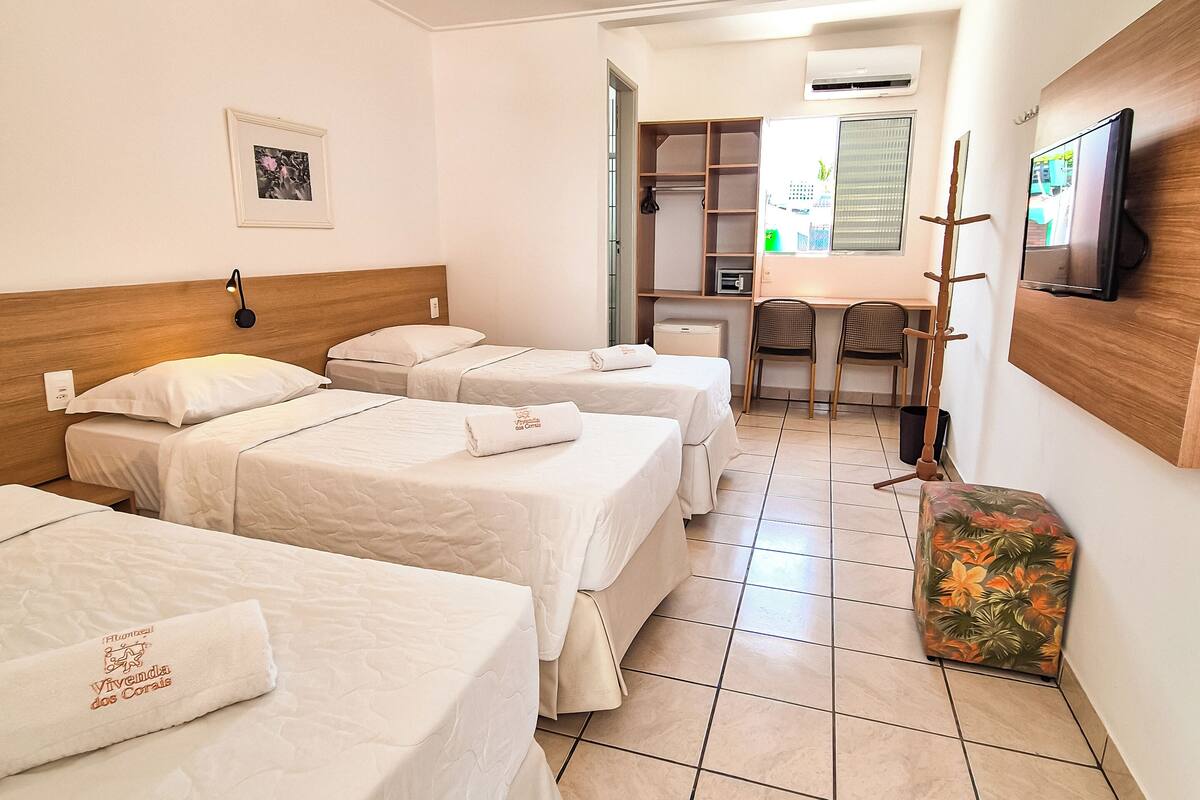 Habitación de hotel con tres camas, un escritorio, un televisor y un baño a la vista a través de una puerta abierta.