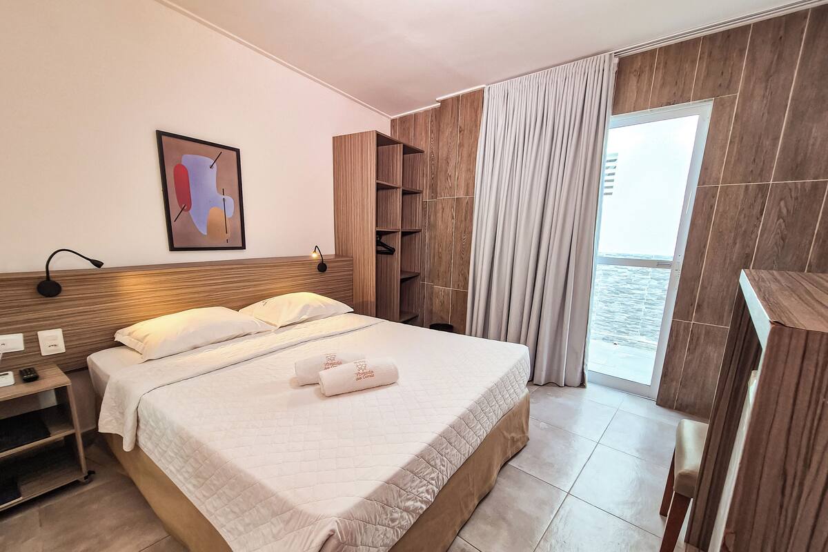 Una habitación de hotel con una cama grande, una mesita de noche, un armario de madera, un cuadro en la pared y vistas al mar a través de la ventana.