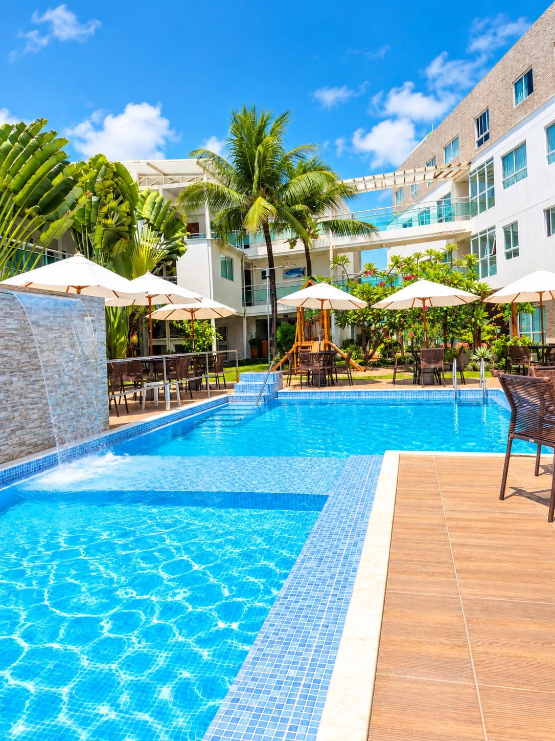 A swimming pool with tiled edges, surrounded by lounge chairs and umbrellas, with a building in the background.