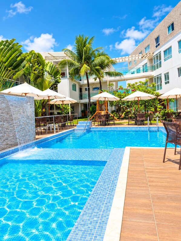 A swimming pool with tiled edges, surrounded by lounge chairs and umbrellas, with a building in the background. - Vivenda dos Corais (Ipojuca)