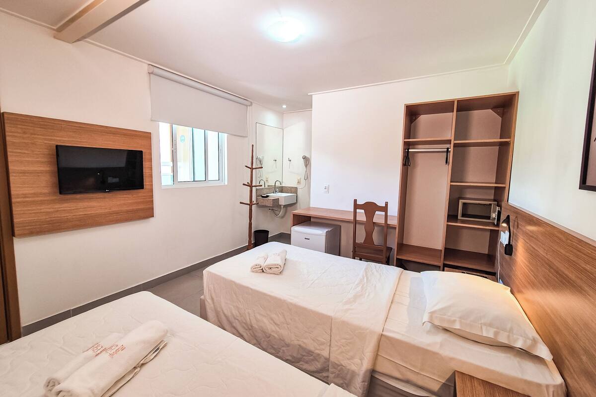 Habitación de hotel con dos camas, televisor montado en la pared, escritorio, silla, armario y ventana.