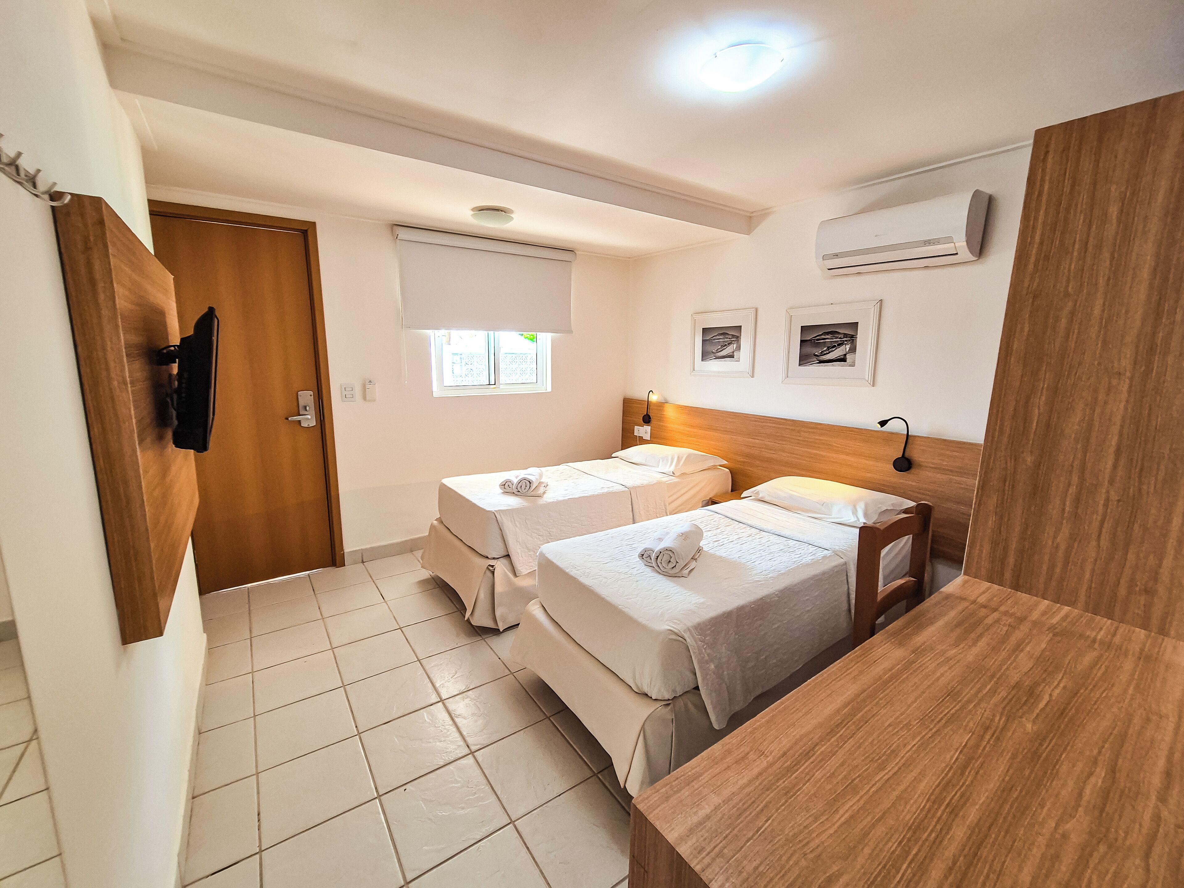 Ein Hotelzimmer mit zwei Einzelbetten, einem Holzkopfteil, einem kleinen Tisch, einem Fenster mit Jalousien, einer Klimaanlage und einer Tür mit Kleiderhaken.
