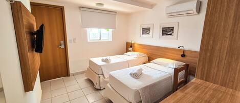Ein Hotelzimmer mit zwei Einzelbetten, einem Holzkopfteil, einem kleinen Tisch, einem Fenster mit Jalousien, einer Klimaanlage und einer Tür mit Kleiderhaken.
