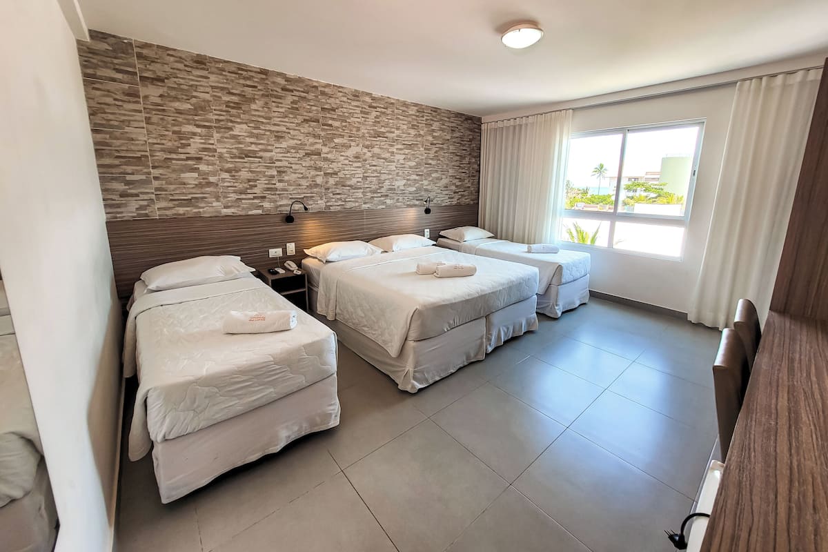 Habitación de hotel con tres camas, pared de piedra, ventana con cortinas y escritorio de madera.
