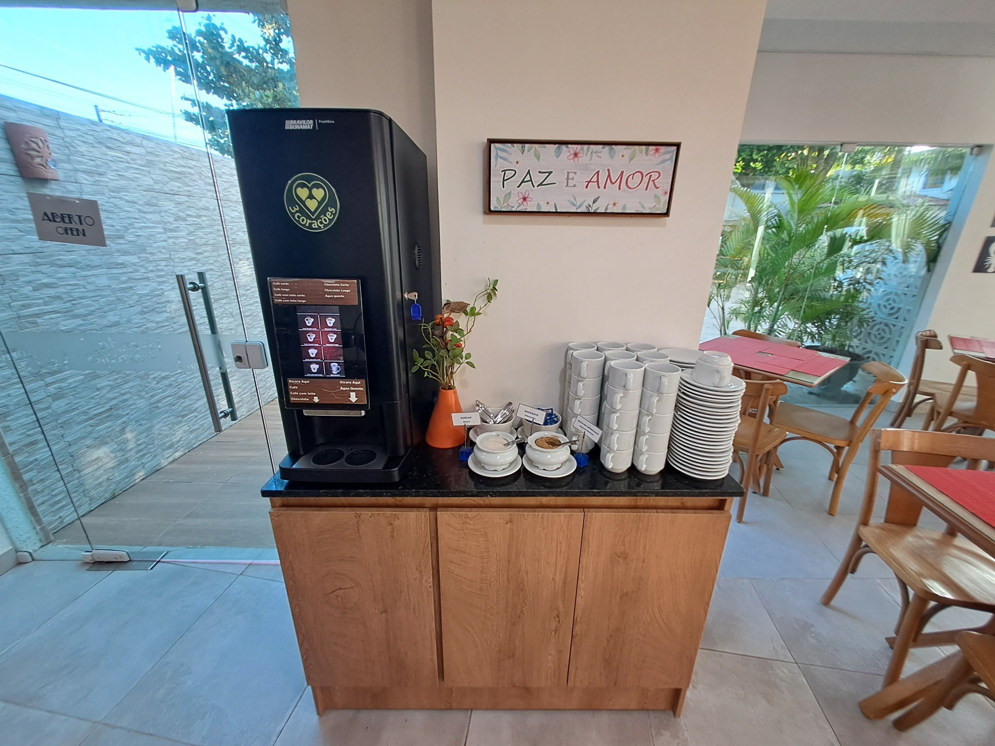 A coffee machine with a menu, a sign reading "ABERTO OPEN", and a wall with "PAZE E AMOR" written on it.