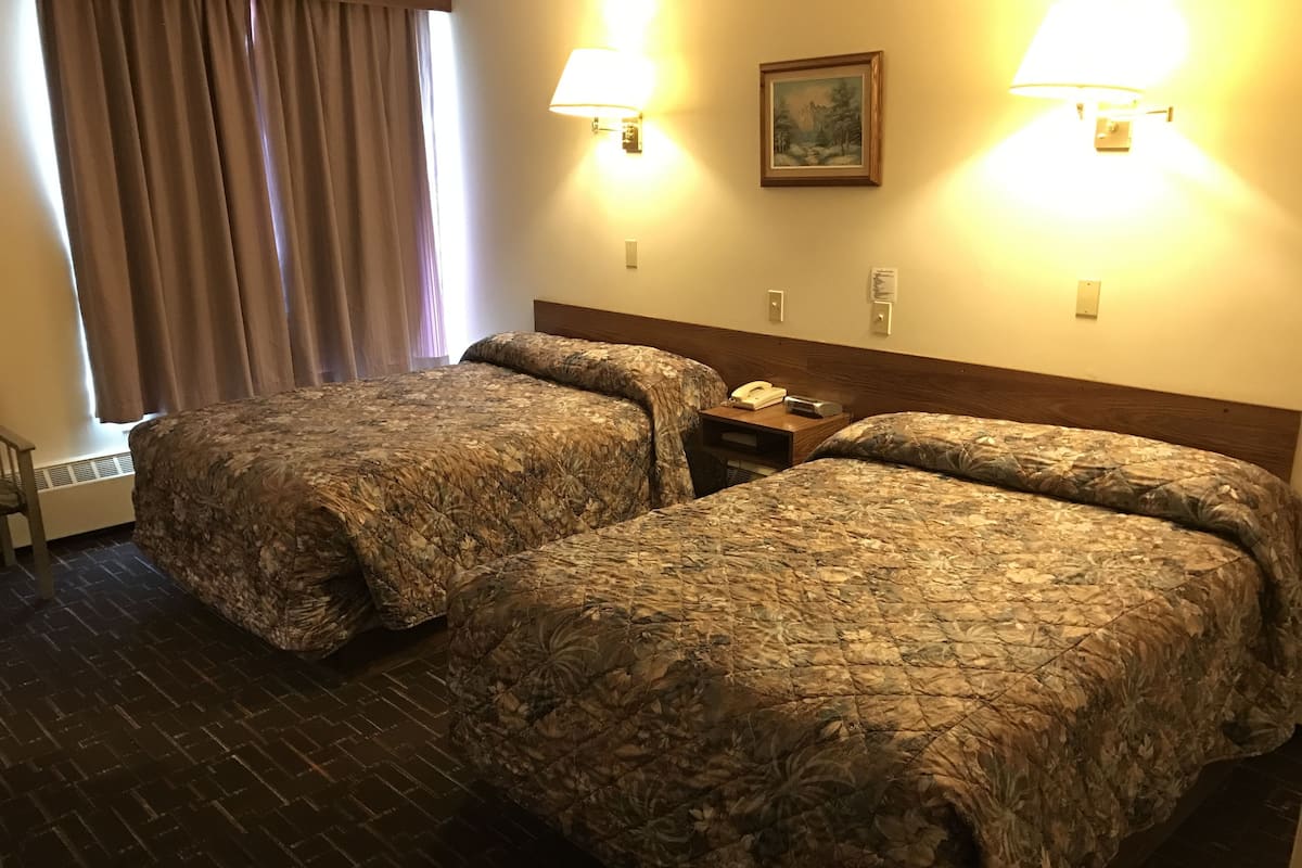 Una habitación de hotel con dos camas, una mesita de noche, una lámpara, un cuadro y una ventana con cortinas.