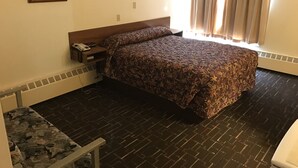 A hotel room with a bed, a nightstand with a phone and remote, a window with curtains, a radiator, and a small table with a chair.