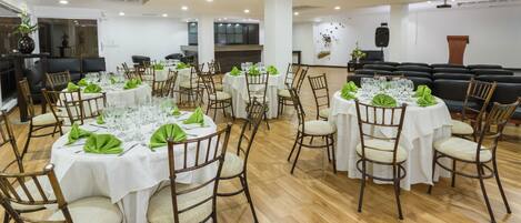 A spacious dining area with multiple round tables, each set with white tablecloths, green napkins, and glassware. The room features wooden flooring and a modern, minimalist design with white walls and ceiling lights.