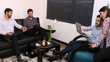 A group of people in a modern lounge area with a large window, a coffee table with a bottle, and a laptop on the couch.