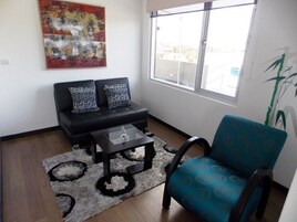 A living room with a black sofa, a blue armchair, a glass coffee table, and a large window with a view of buildings.