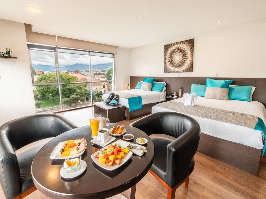 A hotel room with a large window, a bed with multiple pillows, a dining table with breakfast, and a view of the city.