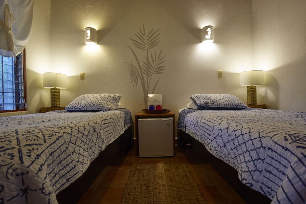 A room with two beds, each with patterned bedding and a small bedside table with a lamp. A refrigerator is placed between the beds.