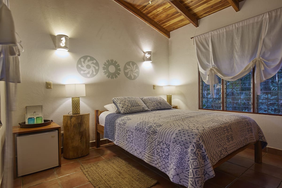 A bedroom with a bed, two bedside lamps, a nightstand, a small refrigerator, and a window with curtains.