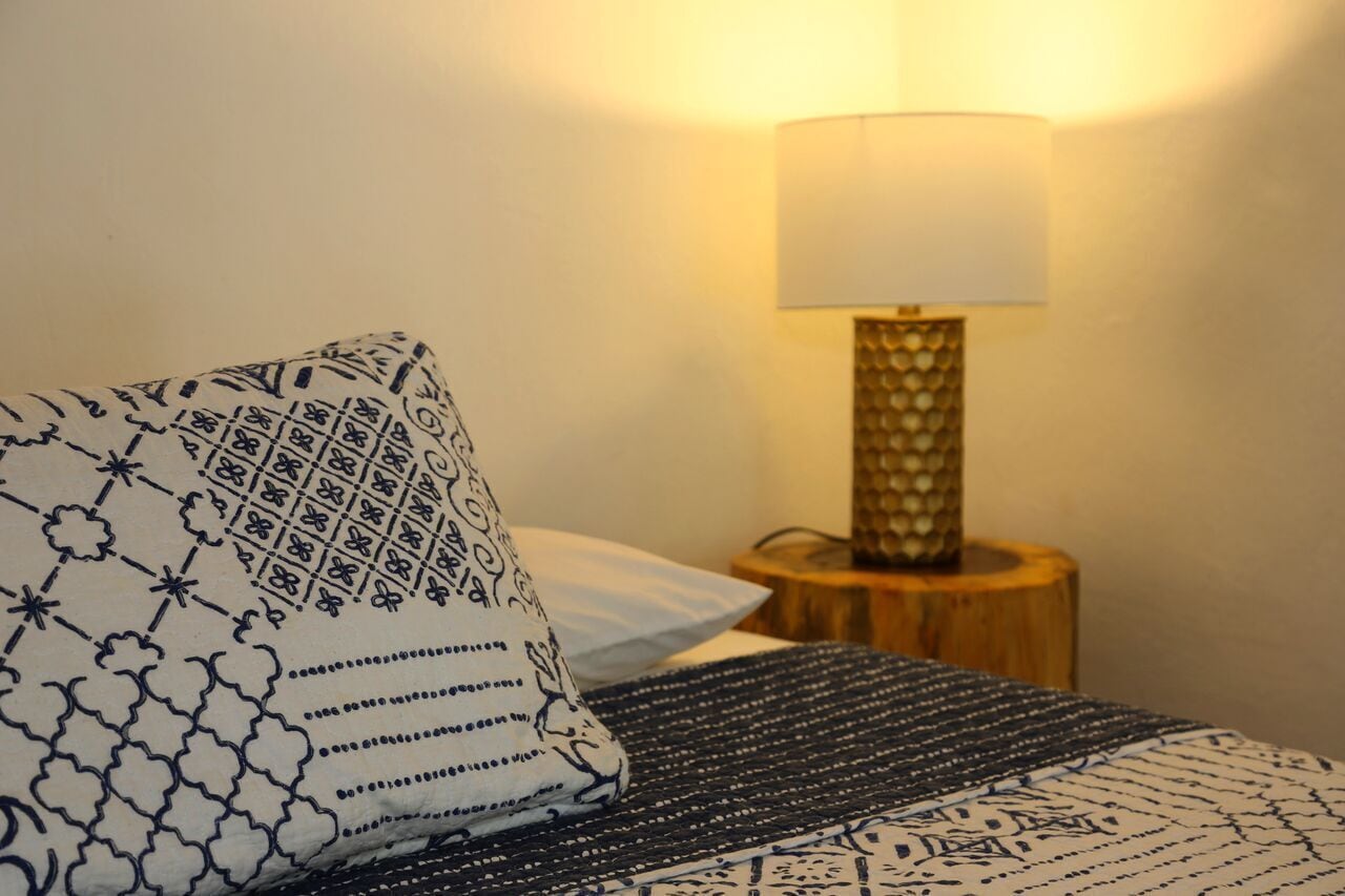 Een net opgemaakt bed met gestreept beddengoed, een bedlampje op een houten nachtkastje en een lamp aan de muur.