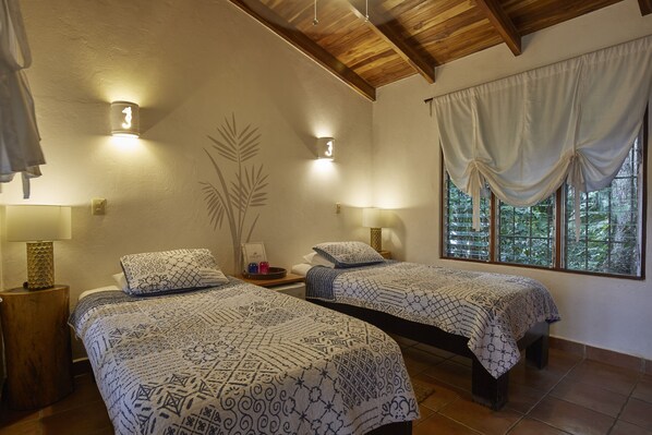 A bedroom with two beds, each with patterned bedding, a wooden ceiling, and a window with curtains.