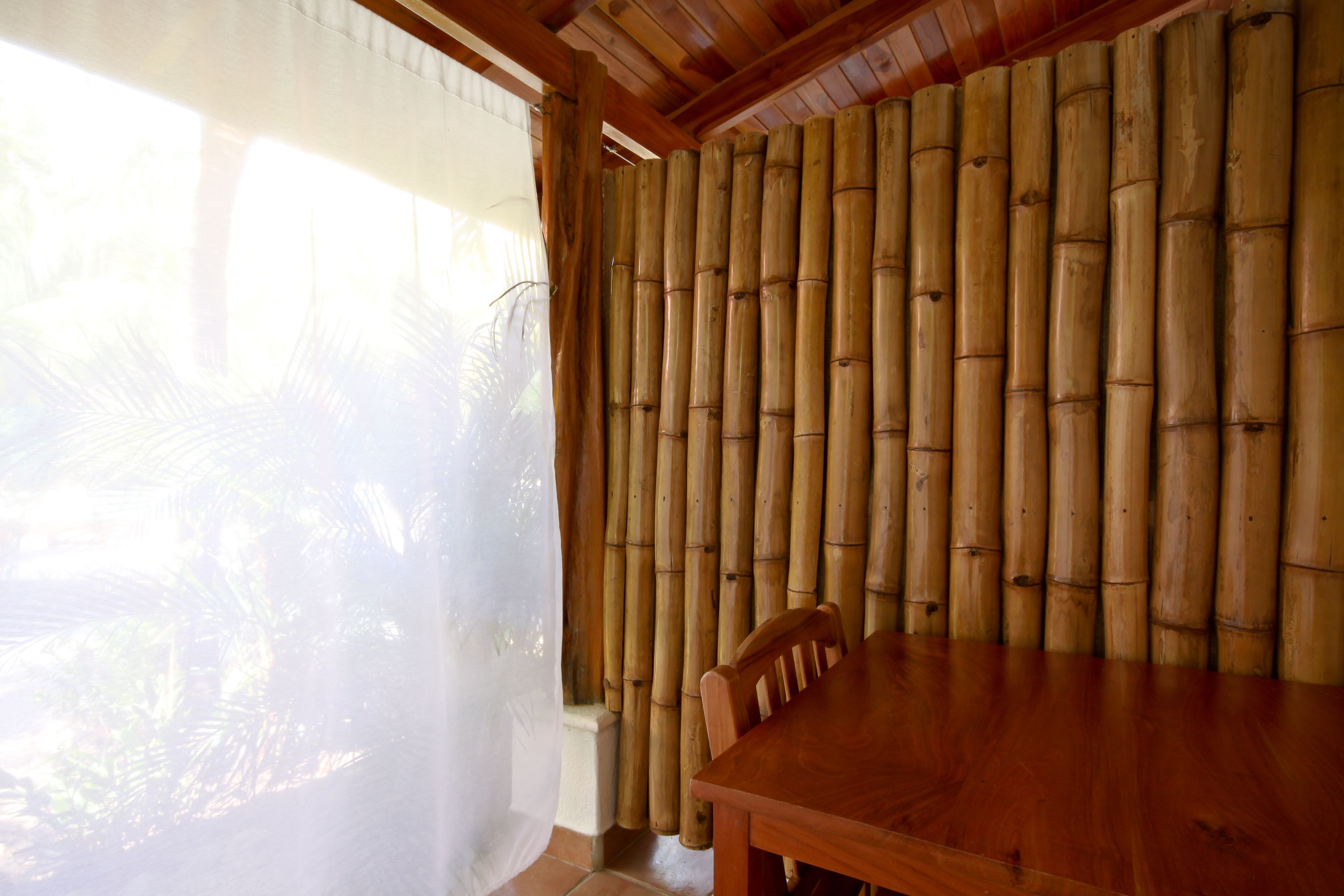 A room with a wooden table and chairs, bamboo wall paneling, and a window with sheer curtains.