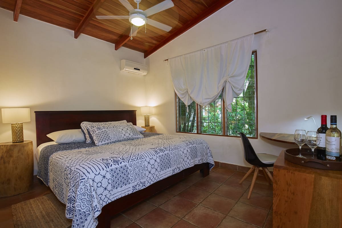 A bedroom with a bed, bedside lamps, a ceiling fan, a window with curtains, a chair, and a small table with wine bottles and glasses.