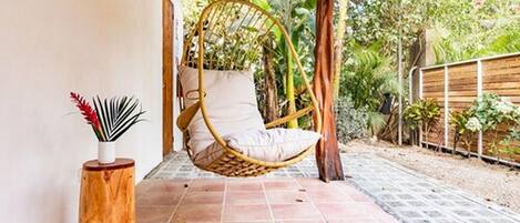 Una silla colgante con cojín, un taburete de madera con una planta, y una zona de patio con baldosas y vegetación al fondo.