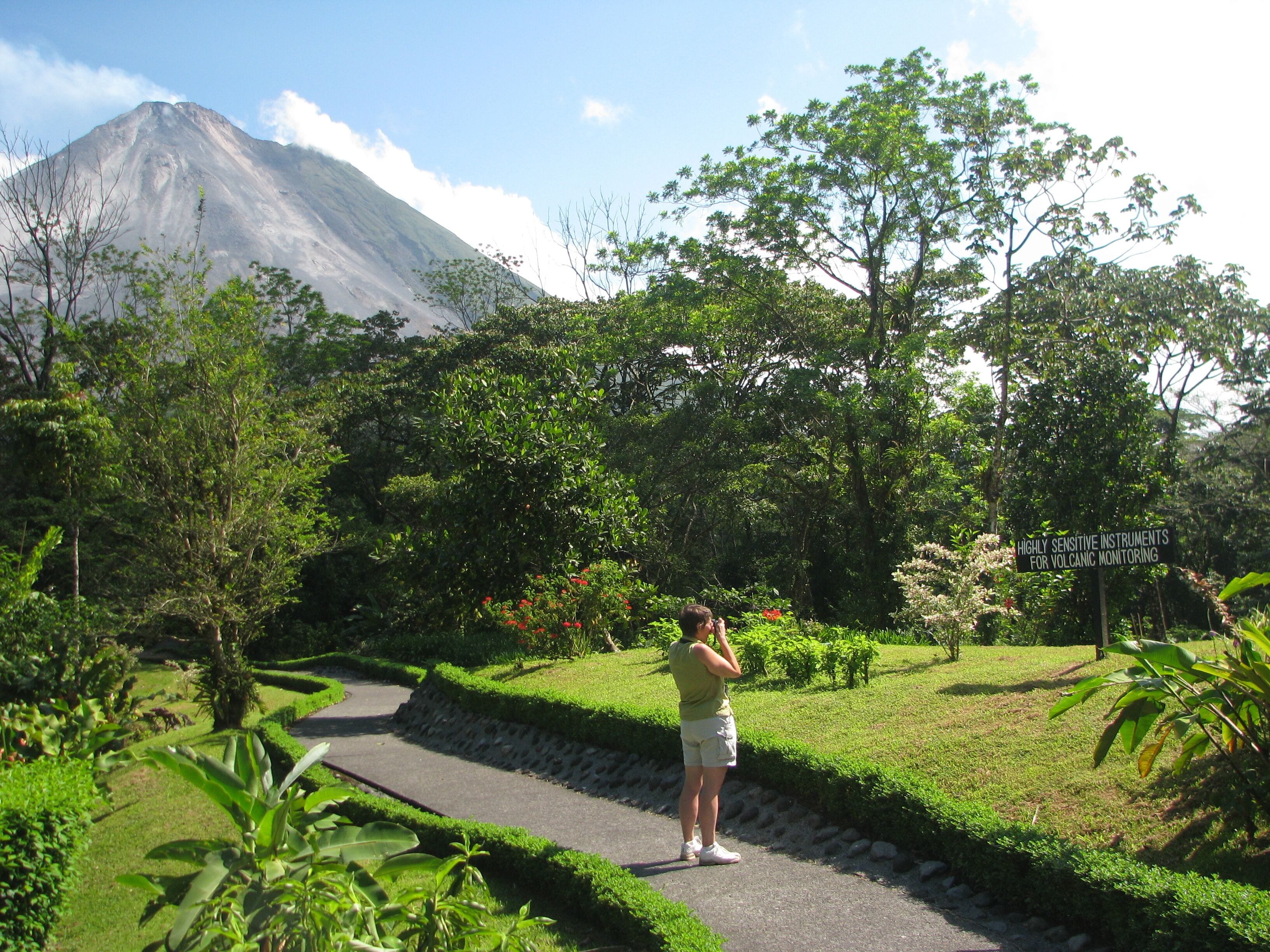 A person taking a photo of a mountain with a sign indicating volcanic monitoring instruments nearby.