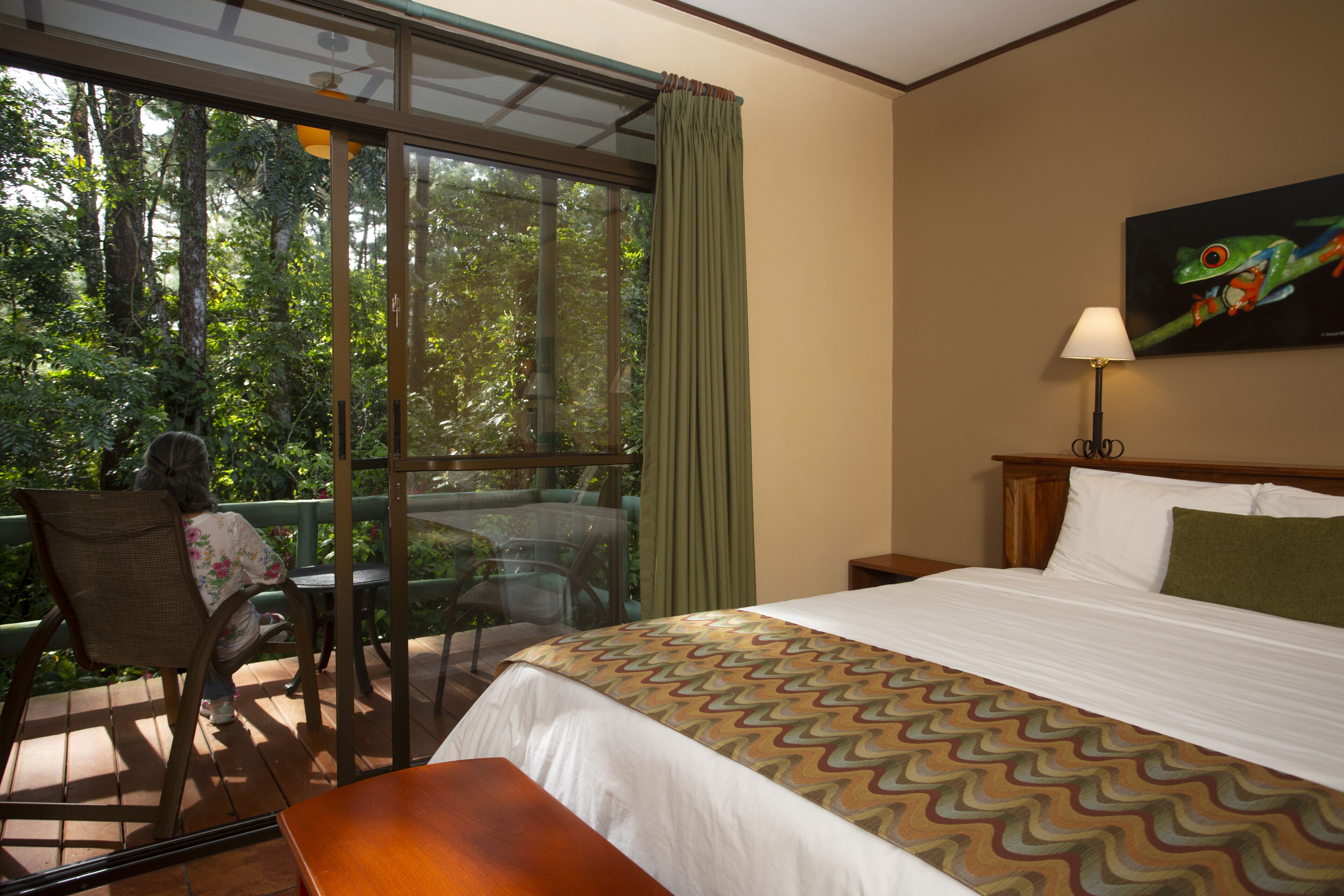 A hotel room with a large window, a bed with a patterned bedspread, a chair, and a table.