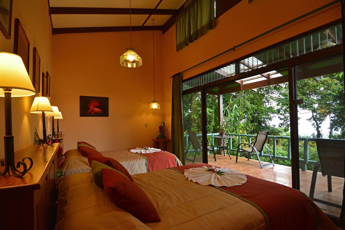 A hotel room with two beds, a balcony with a table and chairs, and a view of greenery outside.