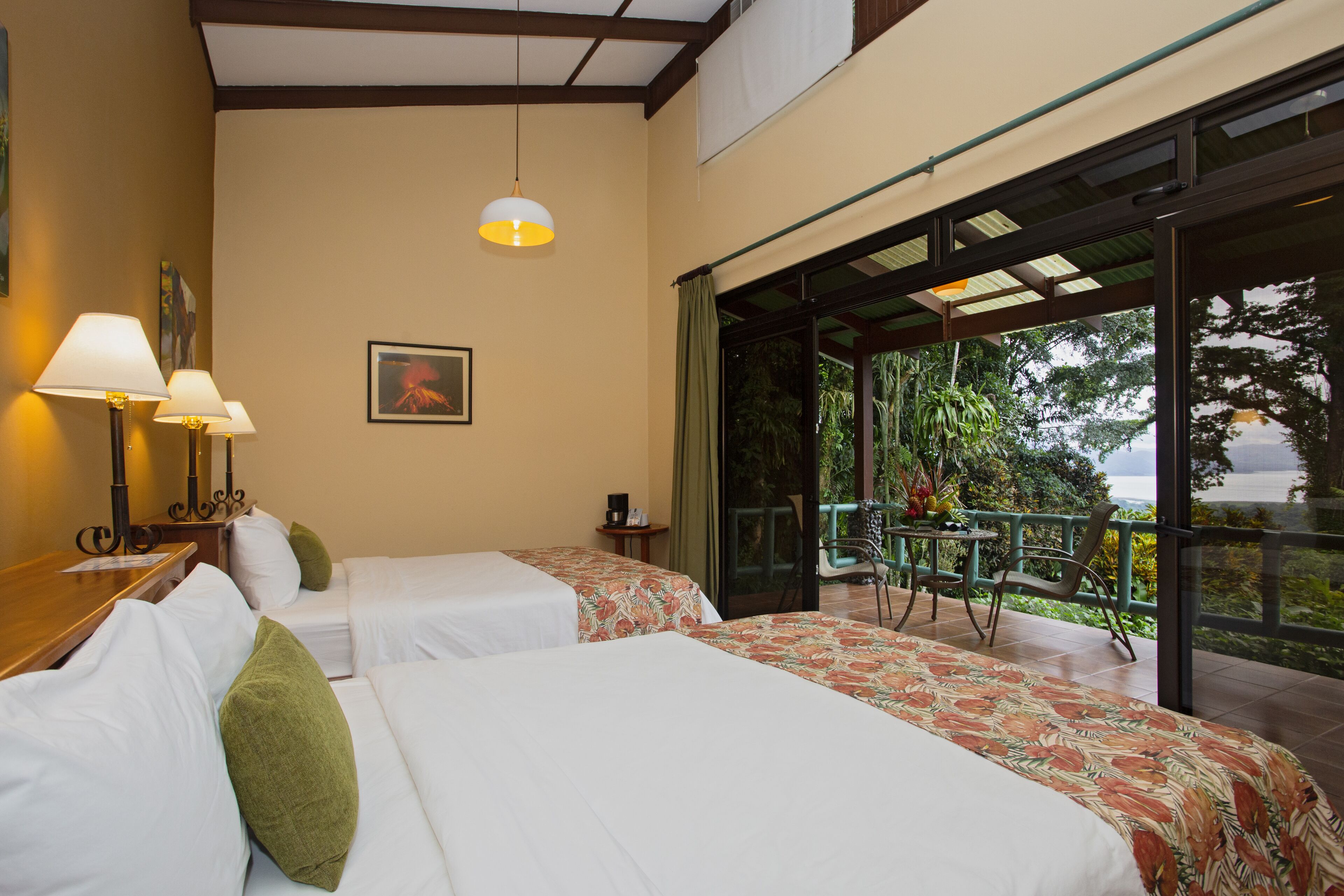A hotel room with two beds, a balcony with a view of greenery, and a table with lamps.