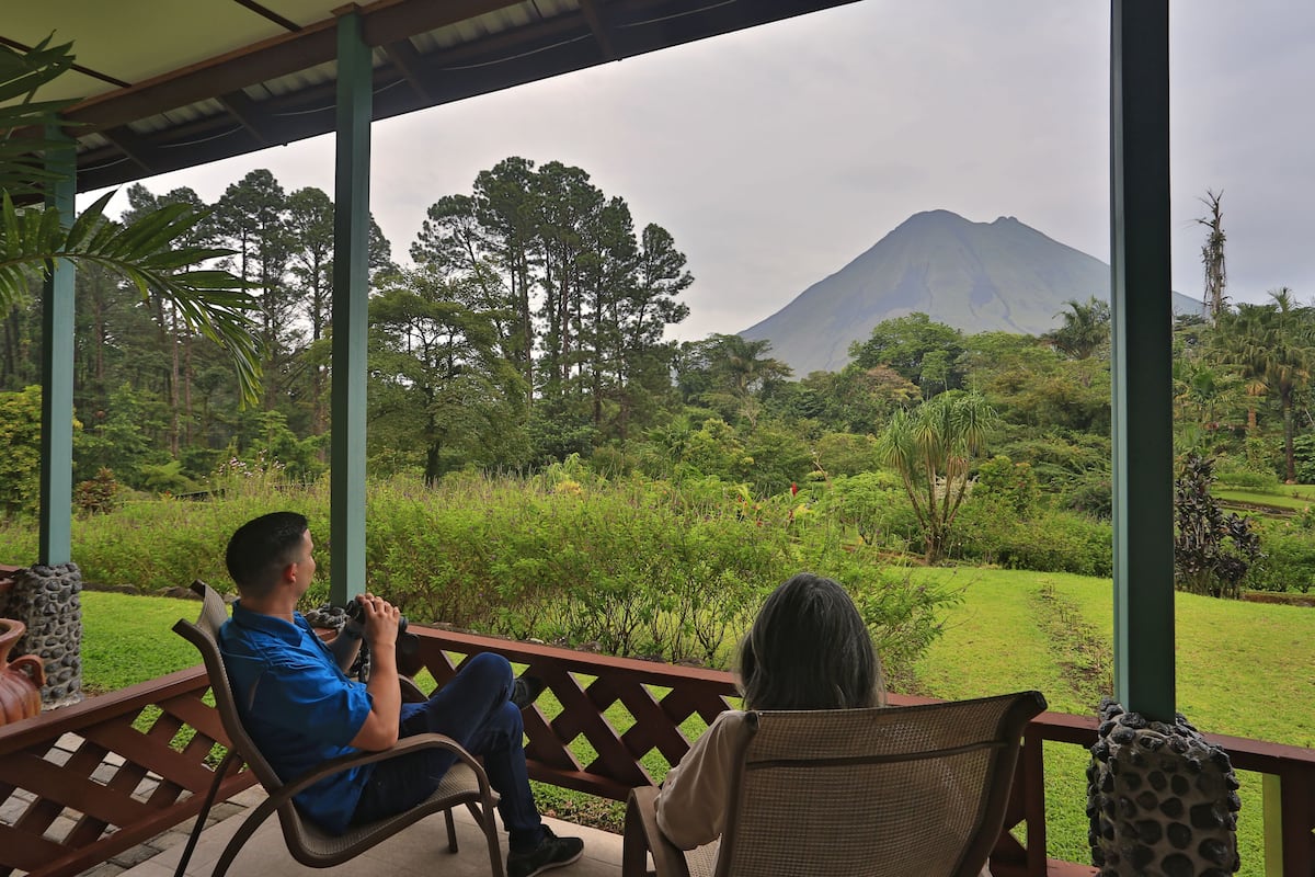 A covered outdoor seating area with two people seated, overlooking a lush landscape with a mountain in the background.