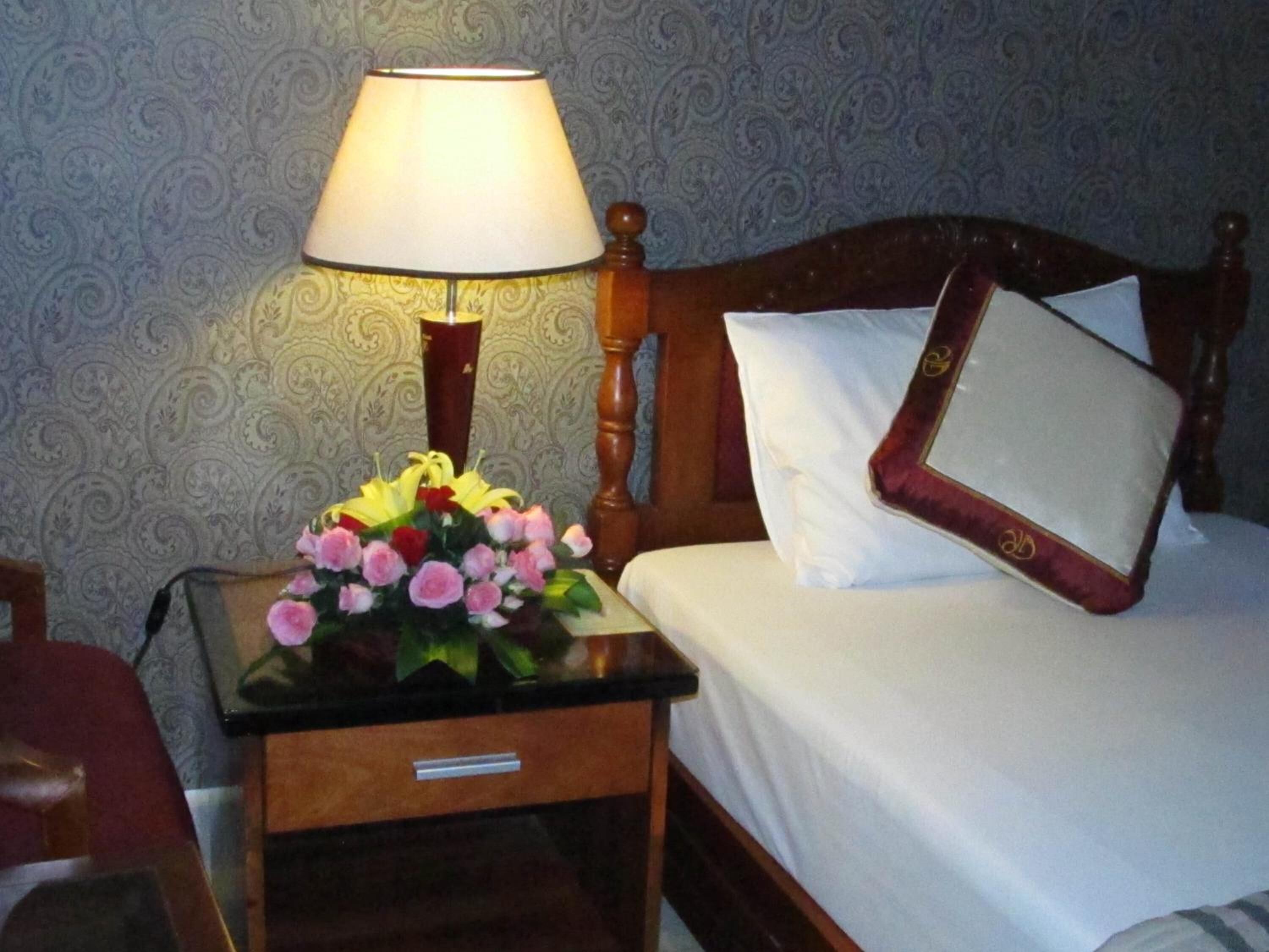 A neatly made bed with white linens, a wooden headboard, and a bedside table with a lamp and a bouquet of flowers.