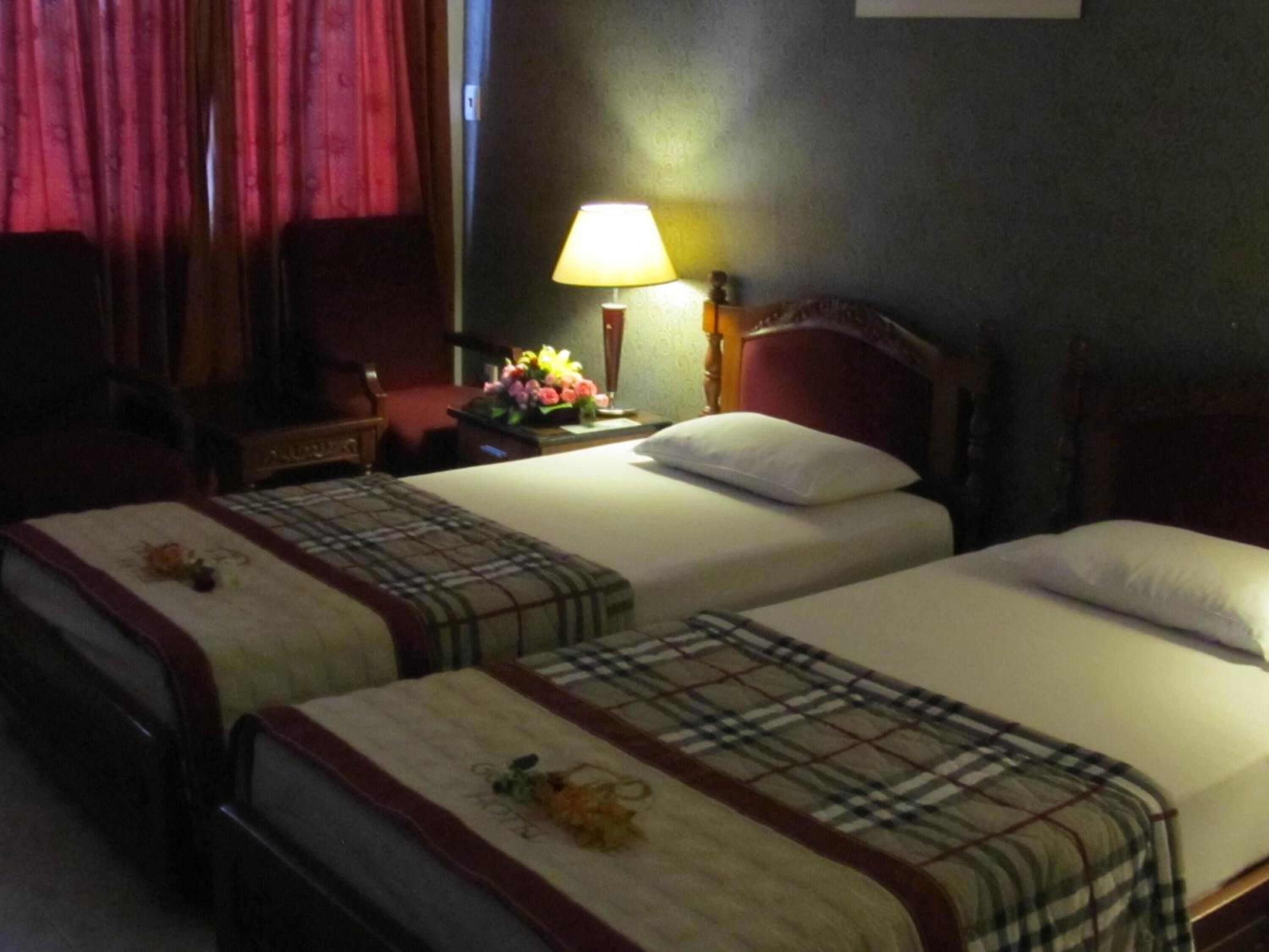 A hotel room with two single beds, each with a plaid bedspread and a decorative flower. There is a nightstand with a lamp and a vase of flowers.