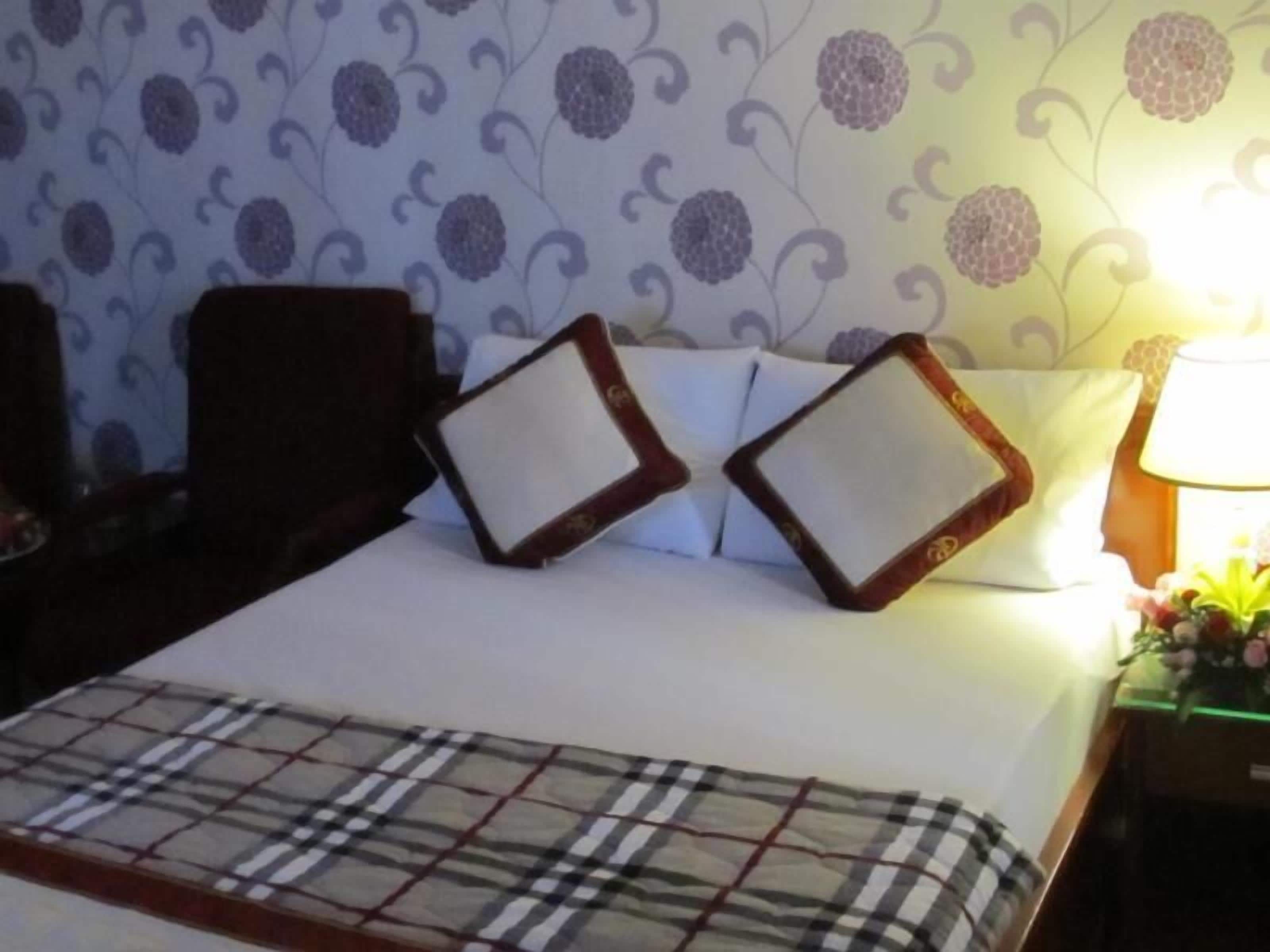 Una cama bien tendida con una colcha escocesa, dos almohadas cuadradas y una mesita de noche con una lámpara y una pequeña planta.