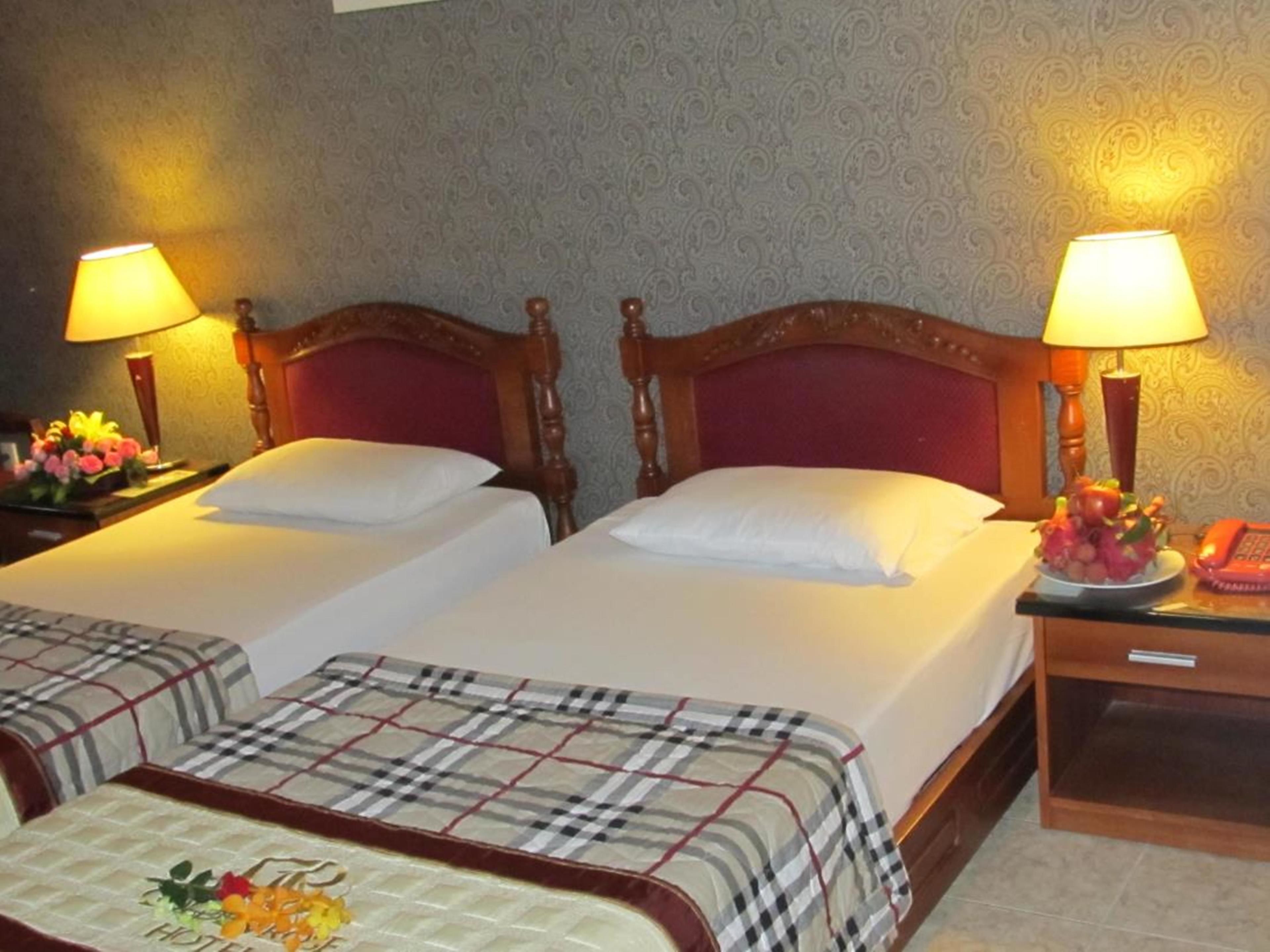 Habitación de hotel con dos camas, cada una con cabecero de madera y ropa de cama blanca. Hay dos mesitas de noche con lámparas y un frutero en una de ellas.