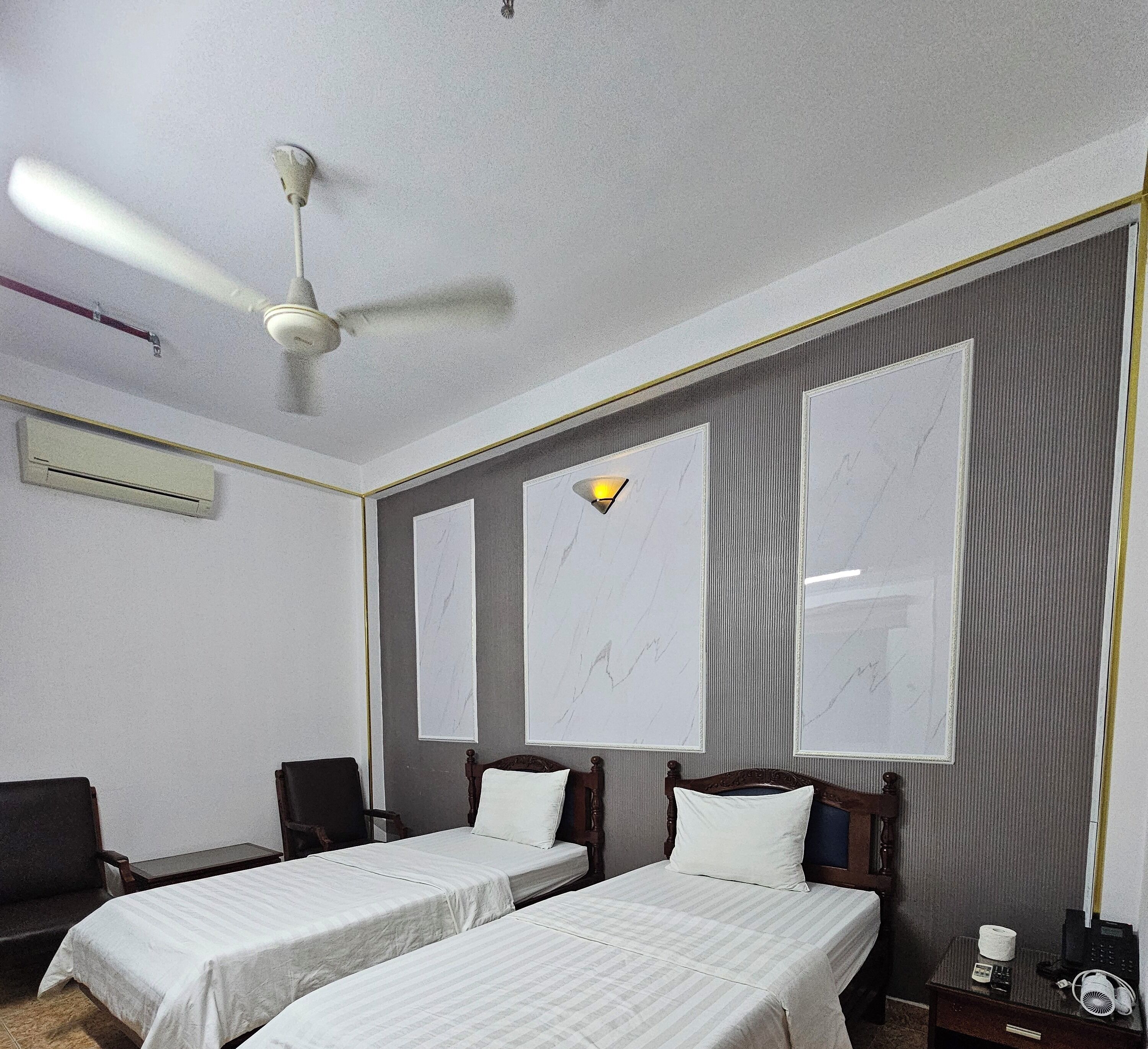 A hotel room with two single beds, a ceiling fan, an air conditioning unit, a bedside table with a phone and a lamp, and large windows with a view of the city.