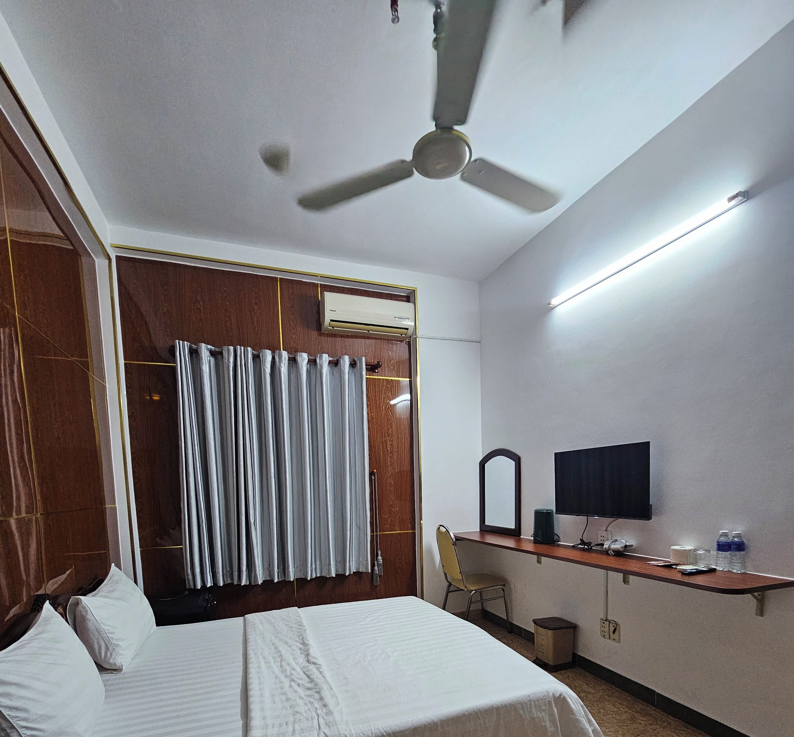 A hotel room with a bed, a desk with a TV, a ceiling fan, and an air conditioning unit.