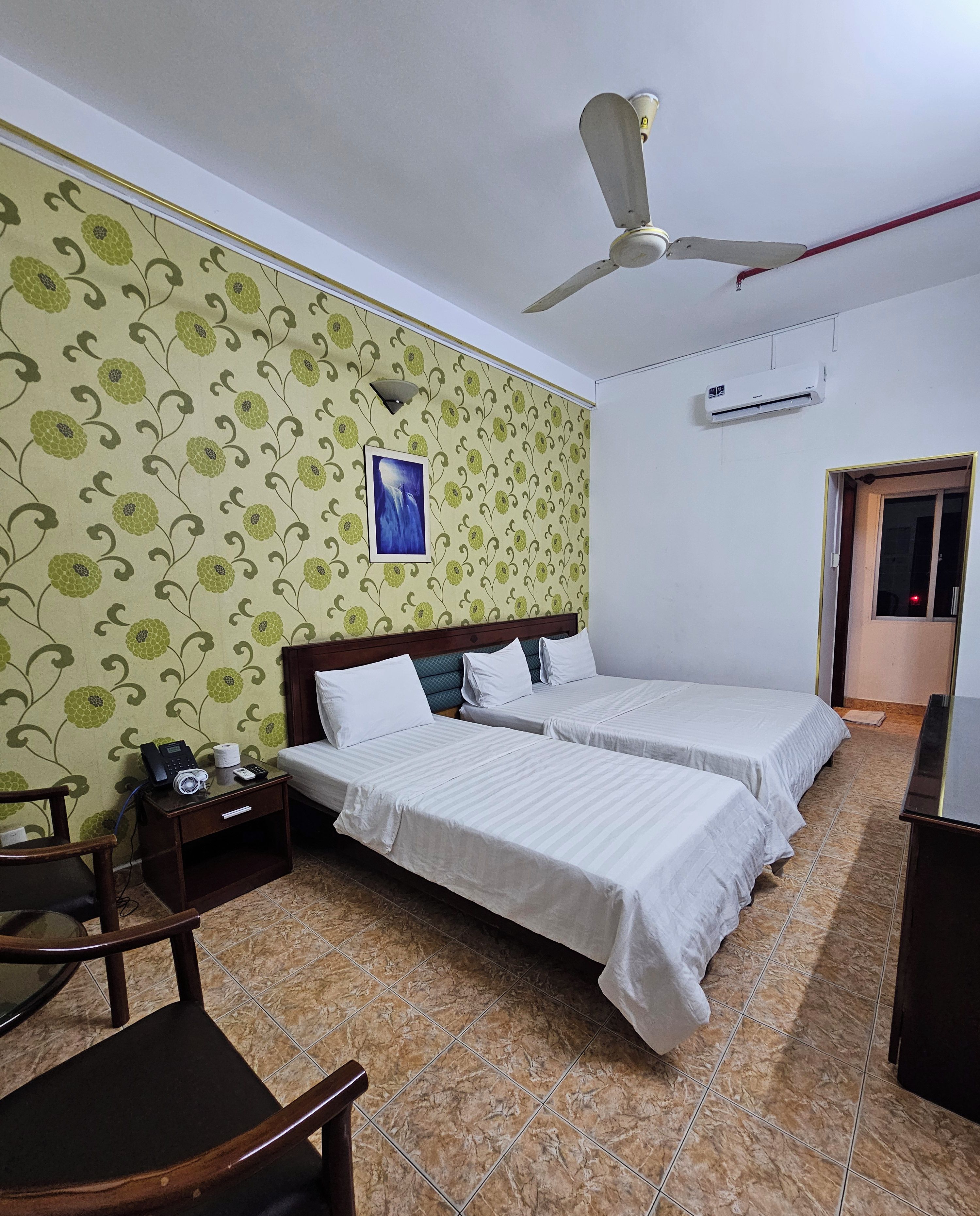 A hotel room with two beds, a wooden nightstand, a chair, a ceiling fan, an air conditioning unit, and a patterned wallpaper.