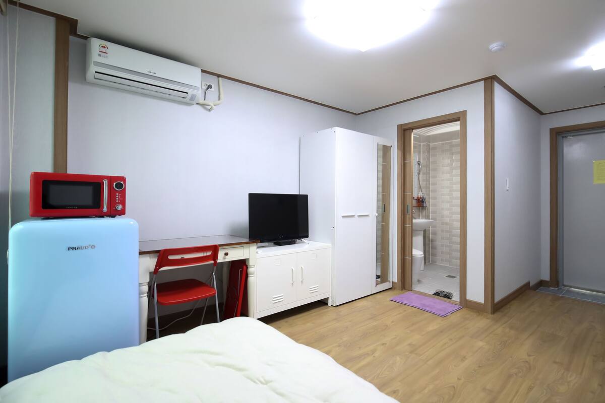 A hotel room with a bed, a desk with a red chair, a television, a white wardrobe, and an air conditioning unit.