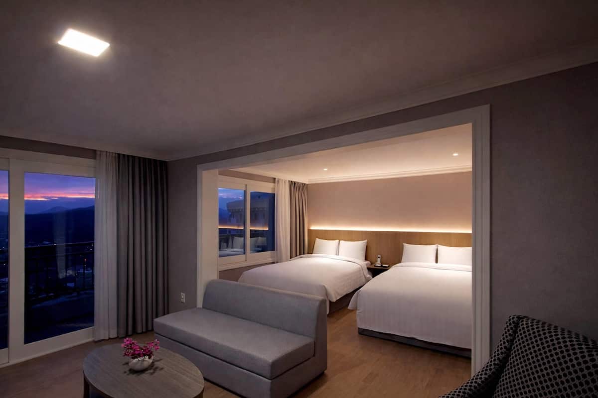 A hotel room with a large bed, a smaller bed, a sofa, a table with flowers, and a view of the city at dusk.