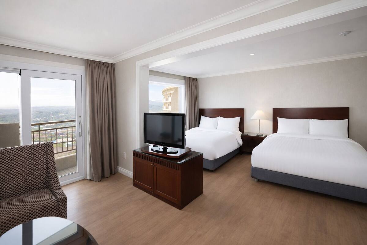 A hotel room with two beds, a TV, a desk, and a balcony with a view.