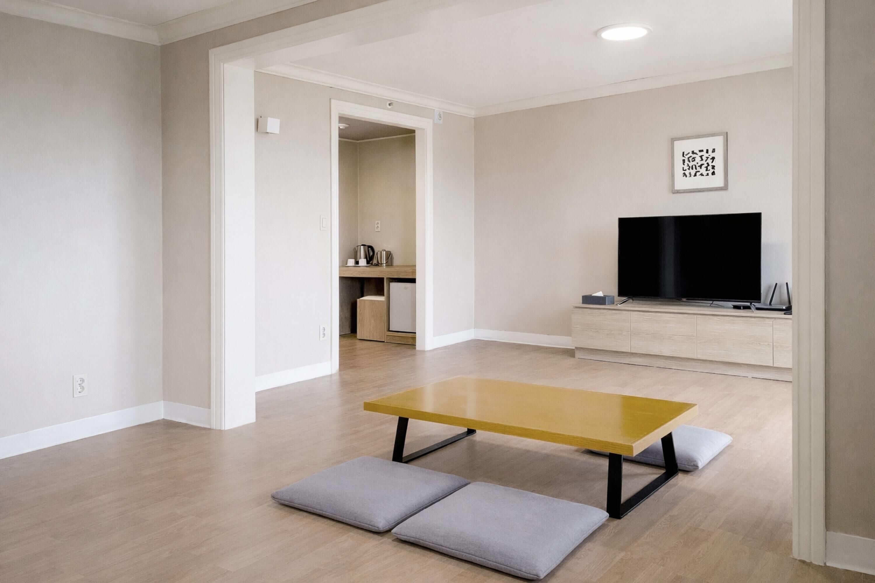 A modern living room with a wooden floor, a yellow table, grey cushions, a flat-screen TV, and a kitchen area in the background.