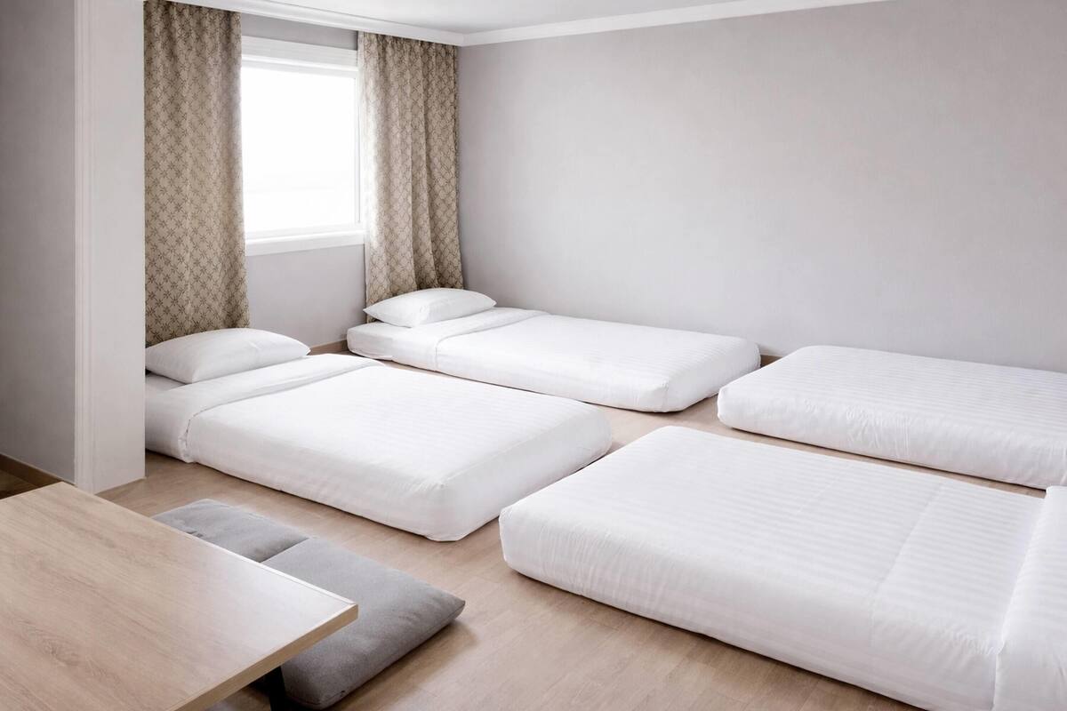 A room with four beds arranged in a corner, a small table, and a window with curtains.