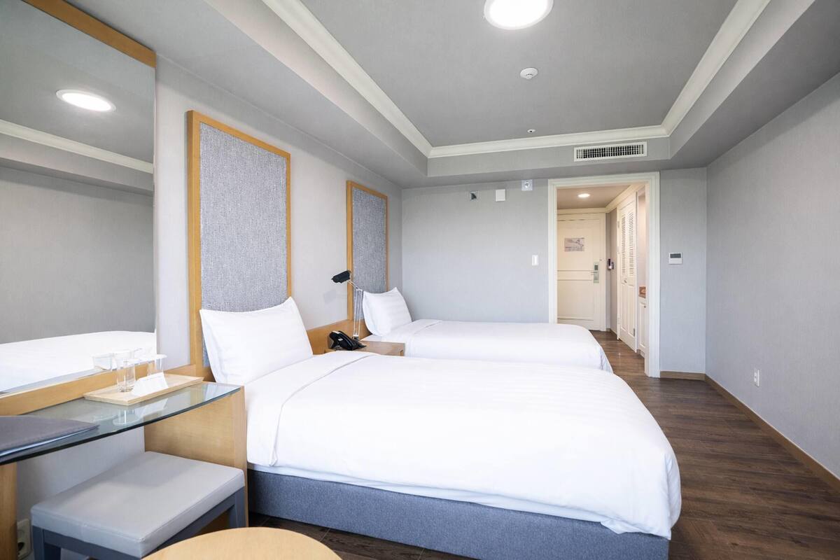 A hotel room with two beds, a desk with a chair, a bedside table, a mirror, and a door leading to another room.