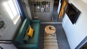 A modern living room with a green sofa, a small round table with a plant, a wall-mounted shelf with decorative items, and a large window with curtains. - The Post Hostel (Jerusalem)