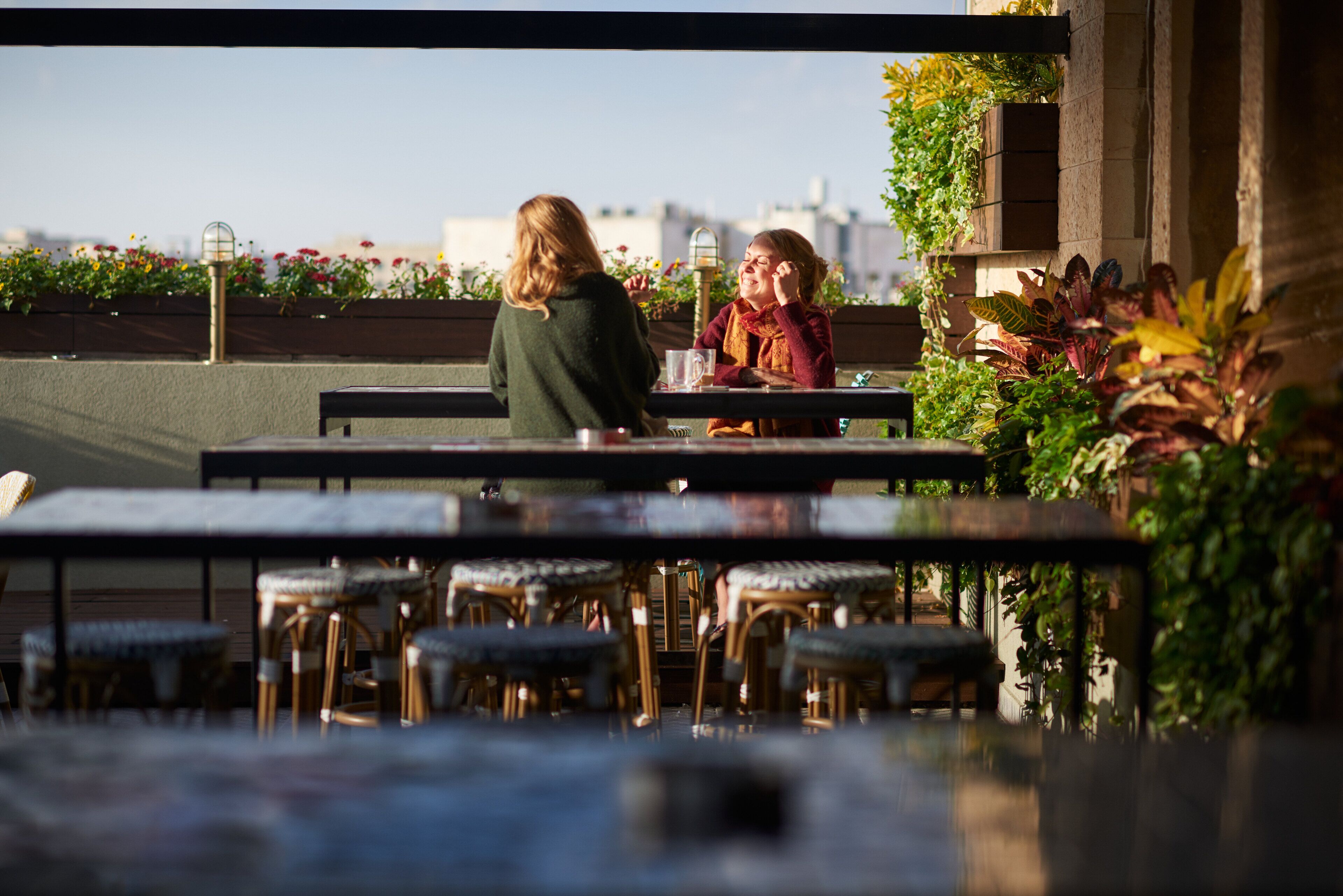 Un área de estar al aire libre con mesas y sillas, rodeada de plantas y vistas a la ciudad.