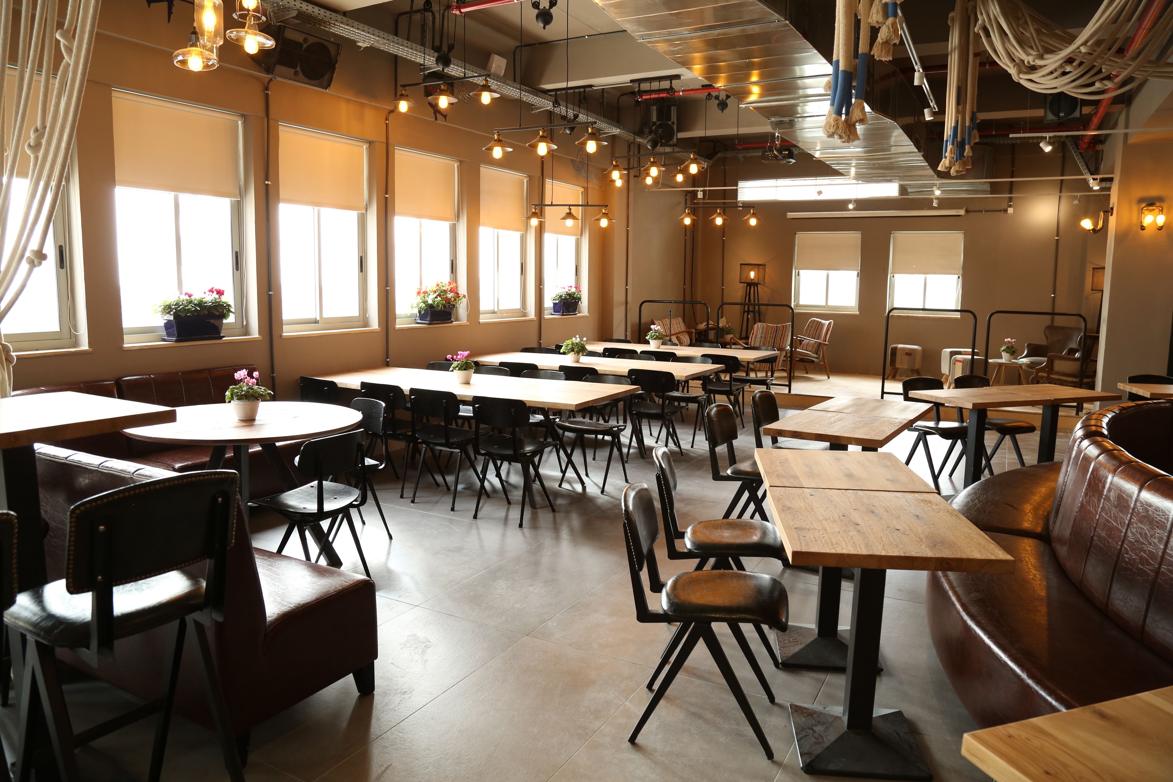 L’intérieur d’un restaurant, avec de nombreuses tables et chaises, de grandes fenêtres équipées de stores et des luminaires suspendus.