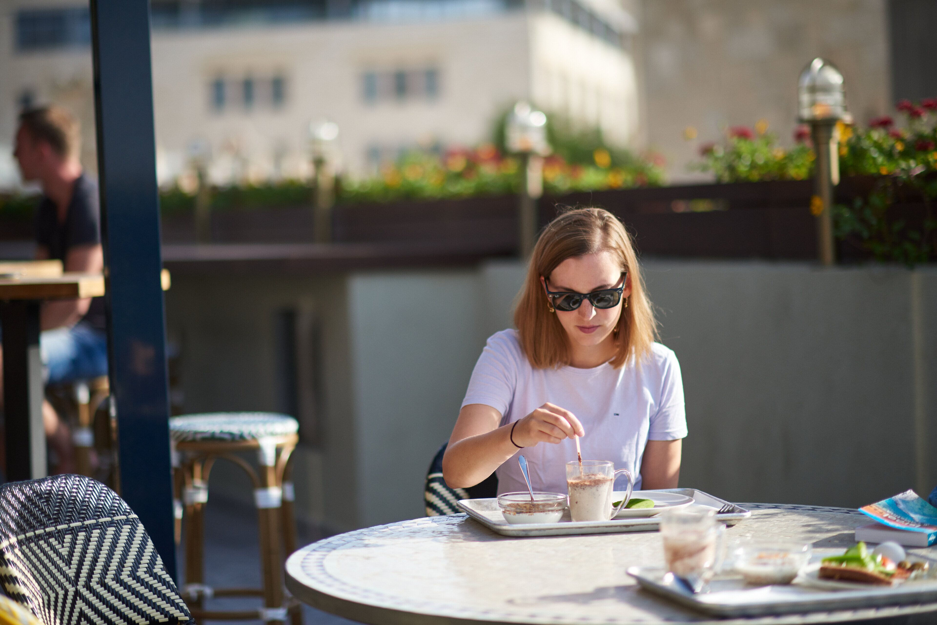 A woman dining at an outdoor cafe with a table set for two, a chair, and a decorative lamp in the background.