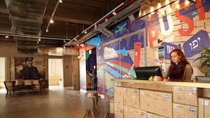 A post office interior with a postal worker at the counter, a mural with postal themes, and stacks of mail on the floor. - The Post Hostel (Jerusalem)