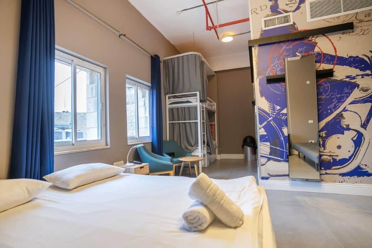 A hotel room with a bed, a chair, a small table, a bunk bed, and a large mirror.