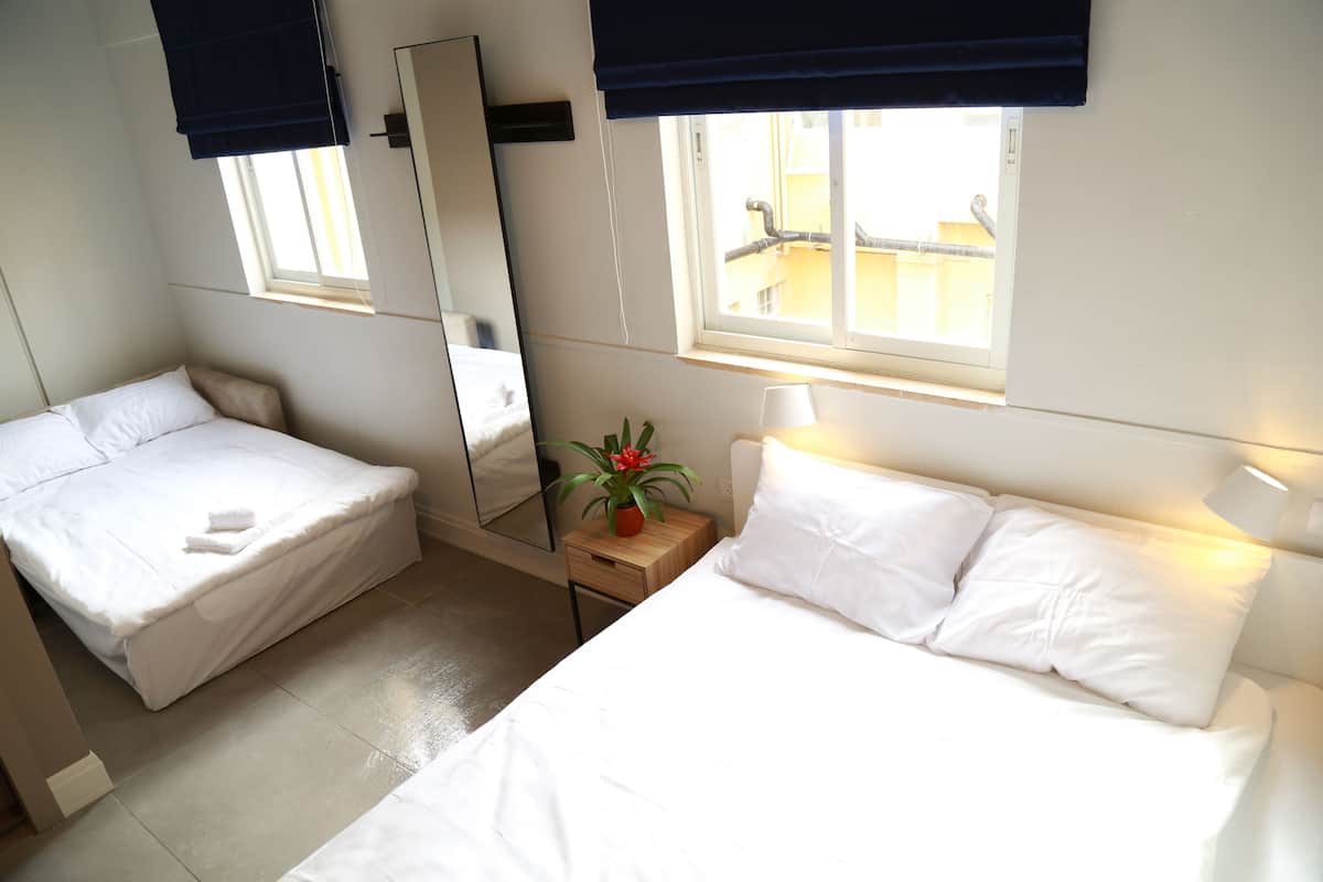 A hotel room with two beds, a mirror, a window with a view of buildings, a small table with a lamp, and a potted plant.